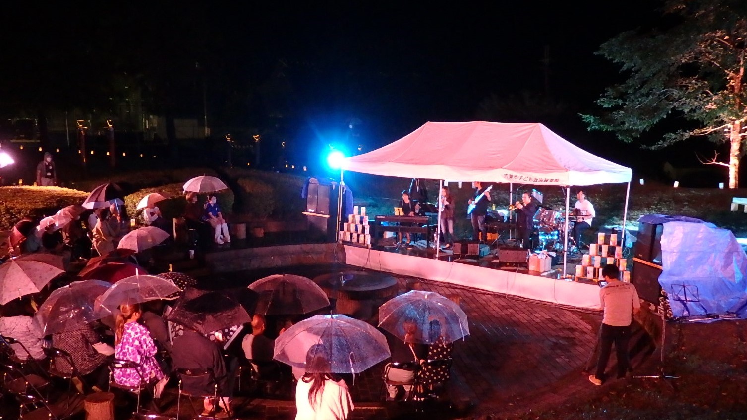 傘を差した来場者らが観月会のステージを囲んで音楽演奏を楽しんでいる写真