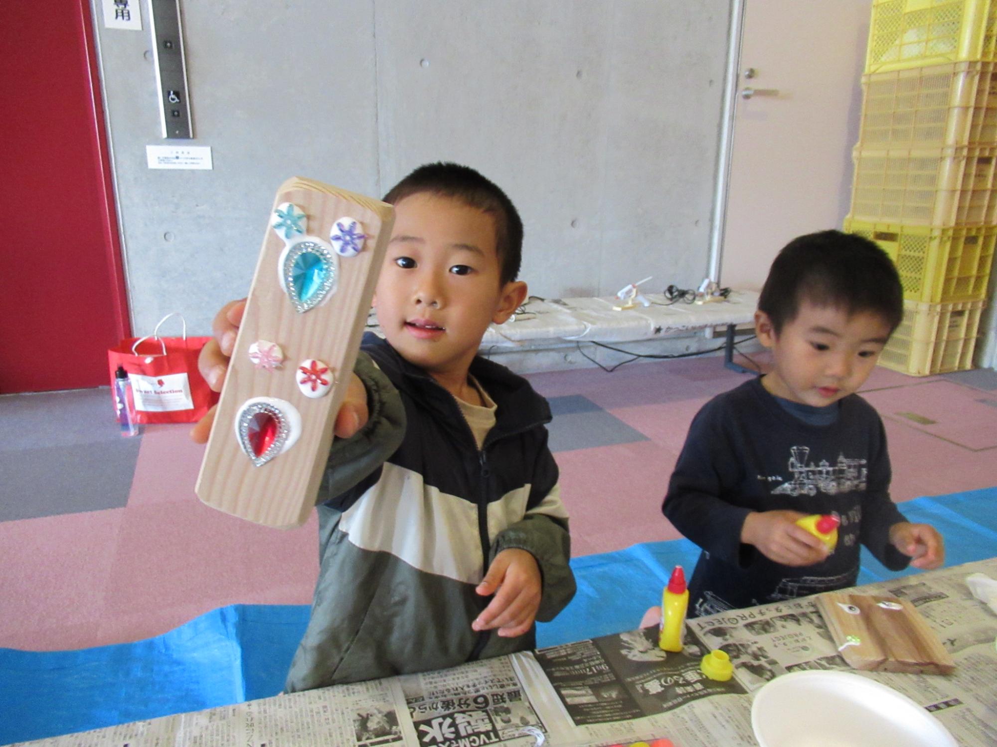 飾り付けたり色を塗ったりした木の板を子どもが見せている写真