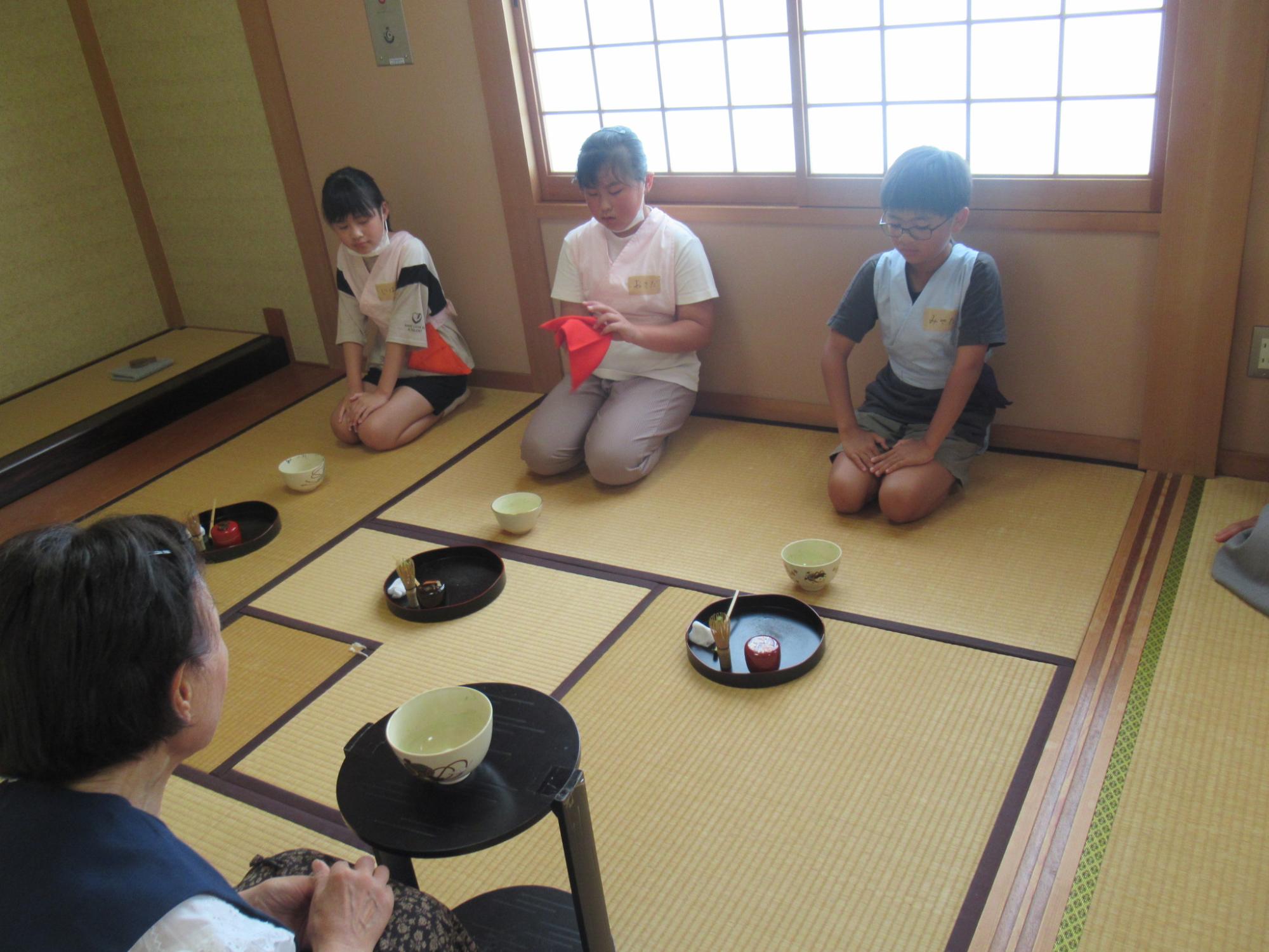 3人の参加者が畳の上に正座した状態で、講師から茶道の作法を聞いている写真