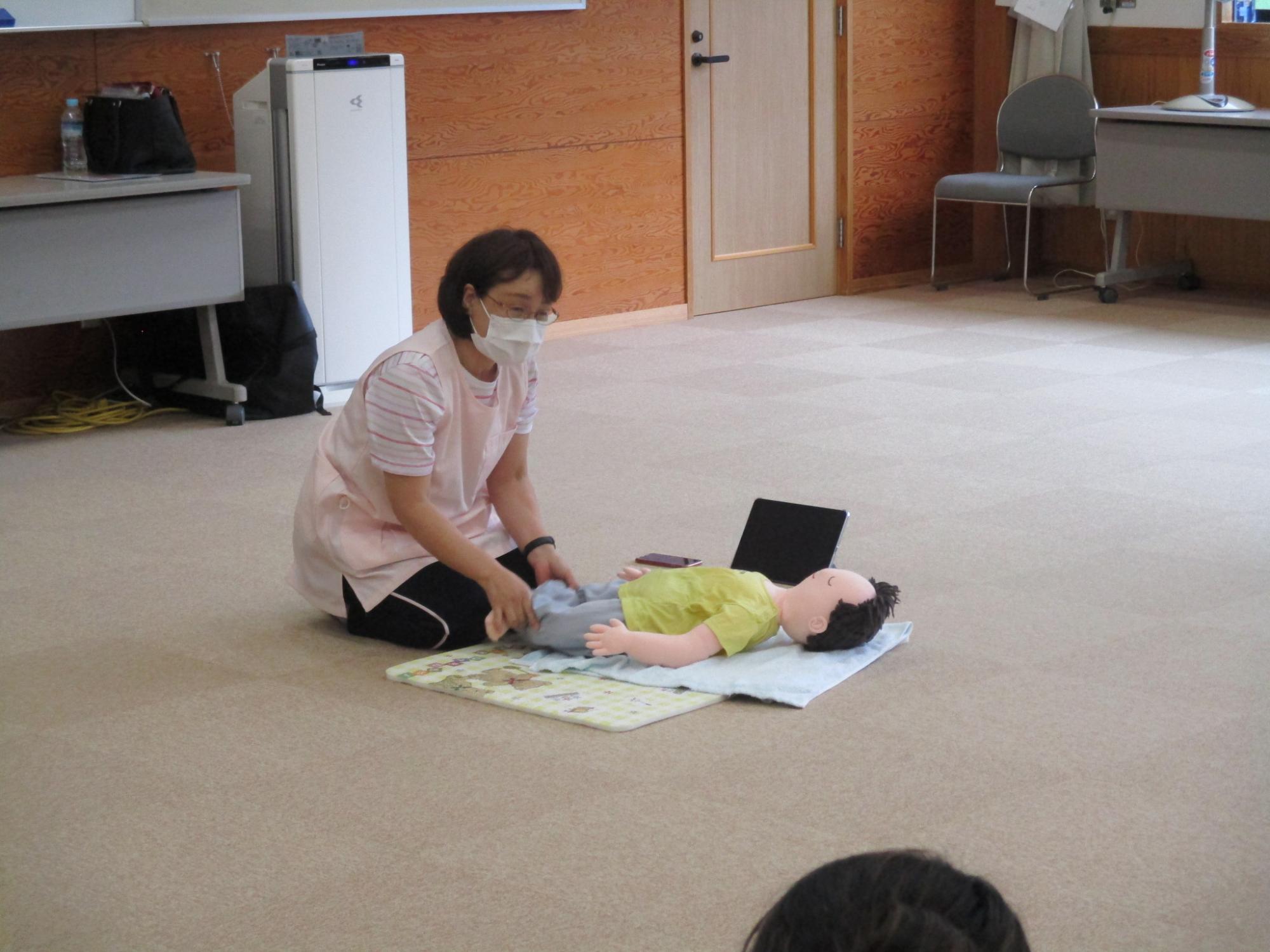講師の八木典子さんが赤ちゃんの人形を見本に参加者にベビーマッサージの方法を教えている写真