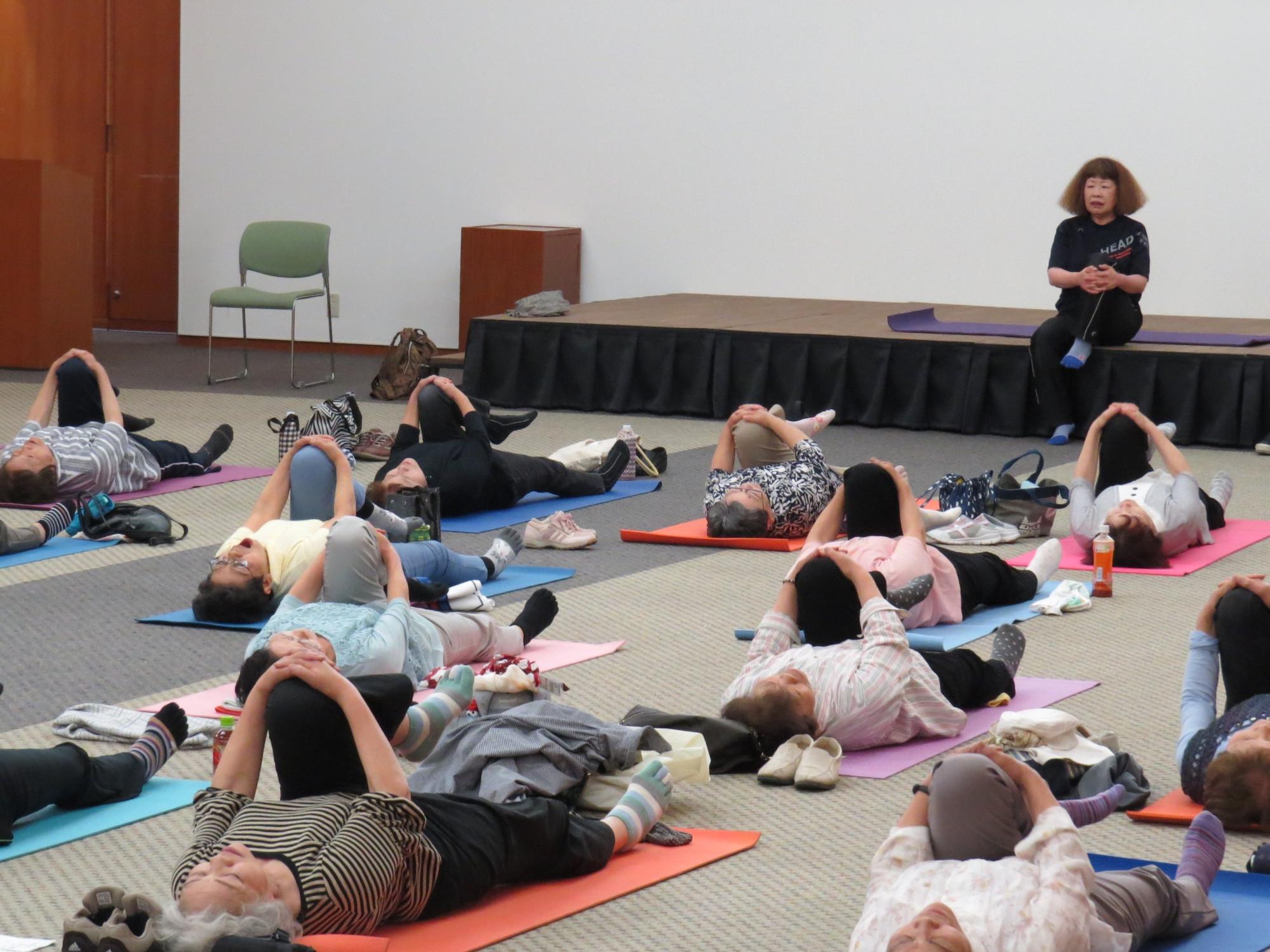 専門講座転ばぬ先の元気塾（健康体操）で床に呼ばマットを引きストレッチを行う大勢の参加者の写真