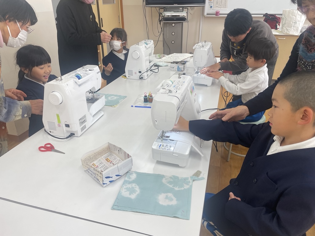大人のサポートを受けながらミシンを使う低学年の子どもたち