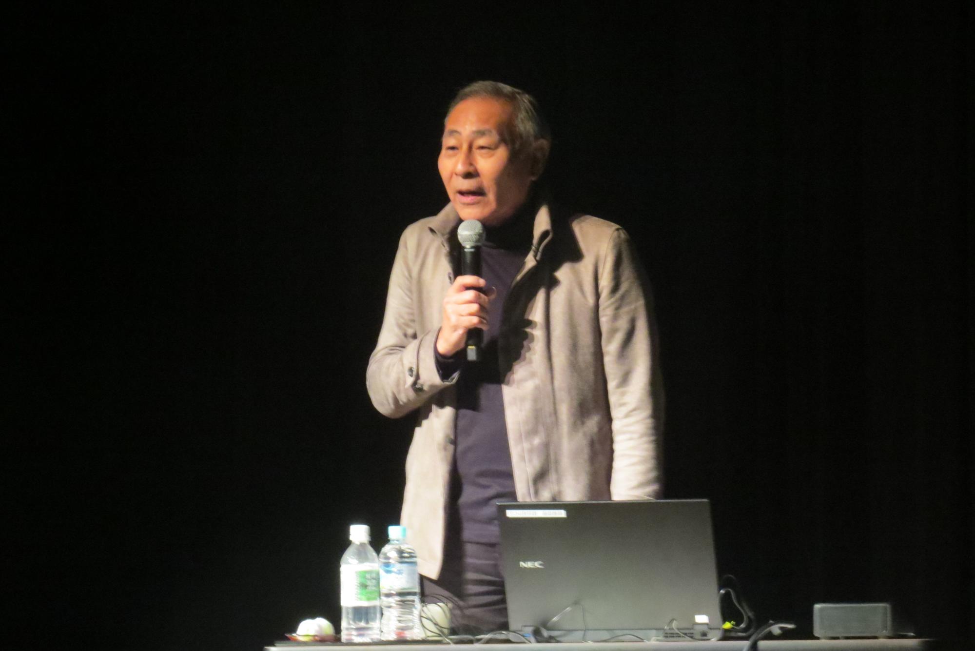 暗い舞台の上でスポットライトに照らされ、夫源病の講演をしている講師の石蔵文信先生の写真