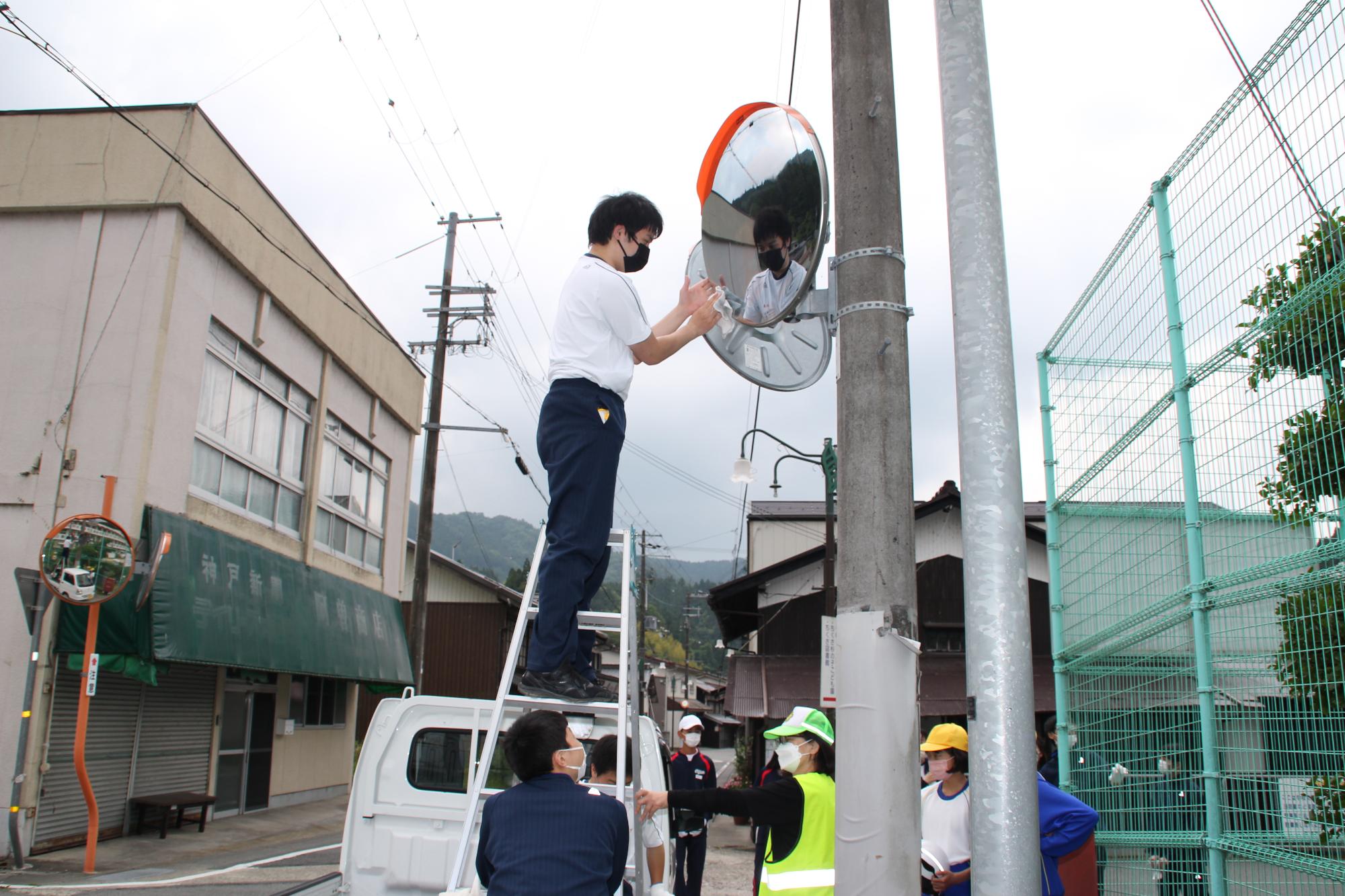 軽トラックの上に脚立を立て、その上で作業をする高校生と、それを見守る小中学生の写真