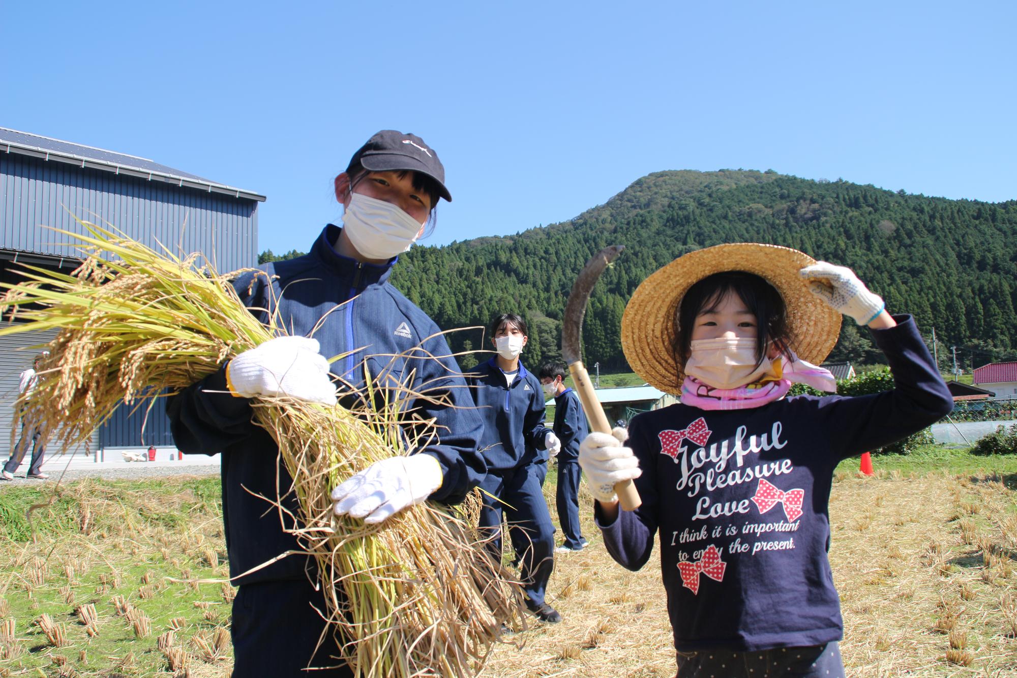 刈り取った稲をもった高校生と鎌を持った小学生が正面を向いて立って並んでいる写真