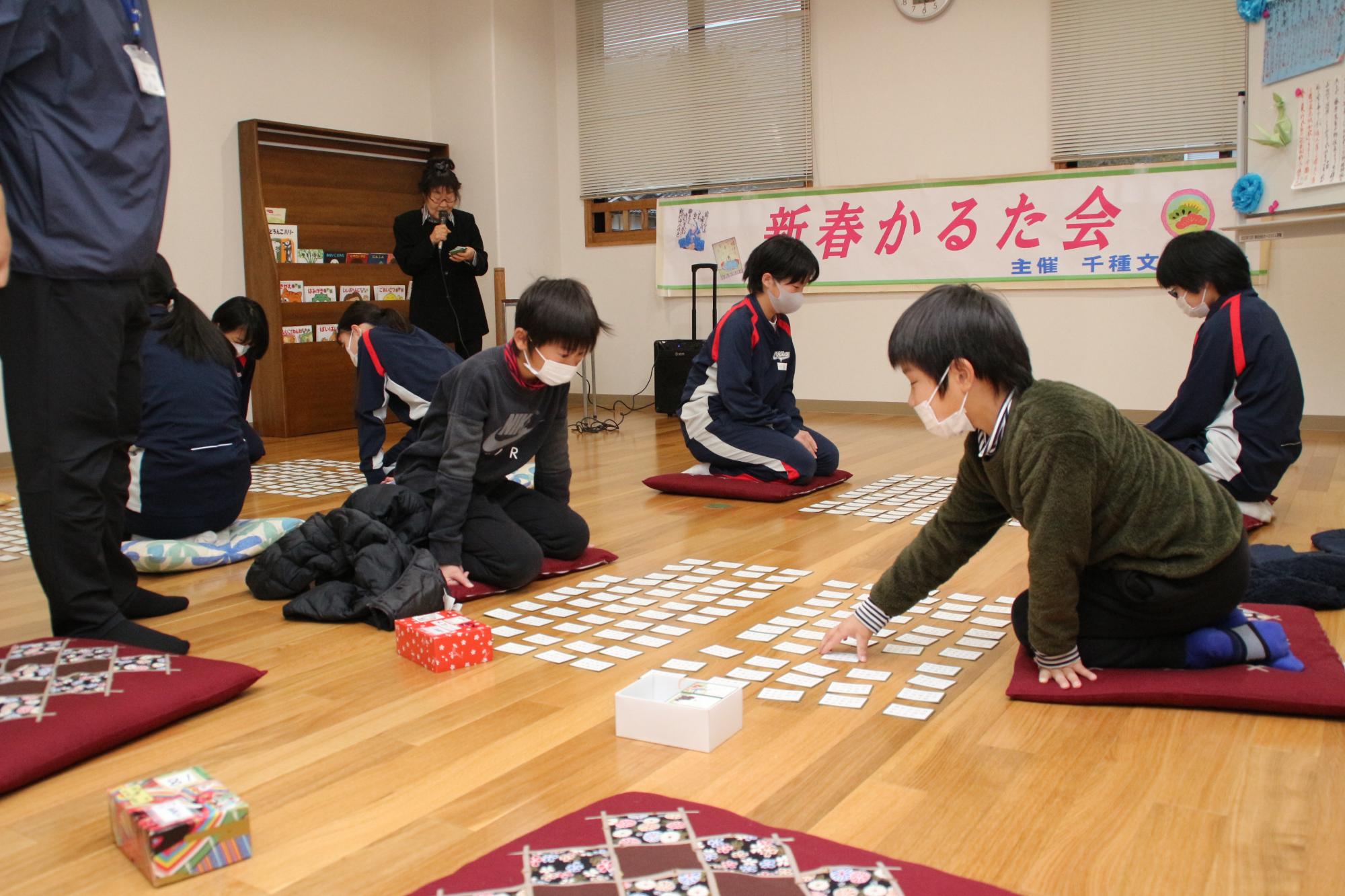 中学生の隣で2人の小学生が向かい合って百人一首で対戦している写真