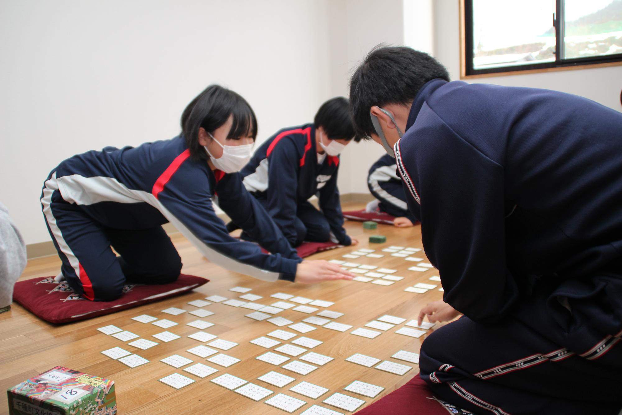 中学生が百人一首を行い、奥にいる女の子が詠まれた句に反応して取ろうとしている様子