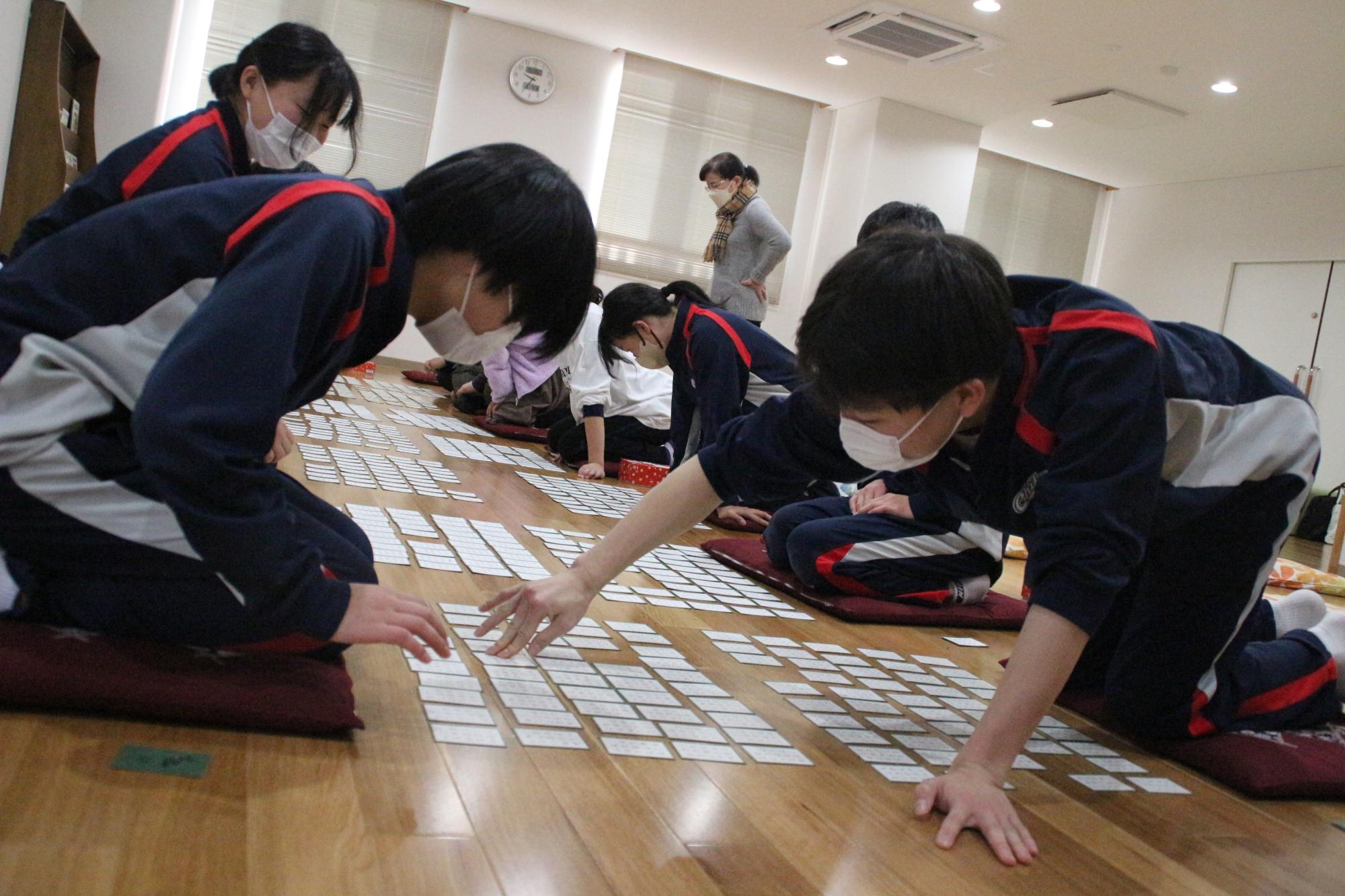 中学生が向かい合わせてに座って、百人一首で詠まれた札を一方の生徒が体を乗り出しながら手で押さえている写真