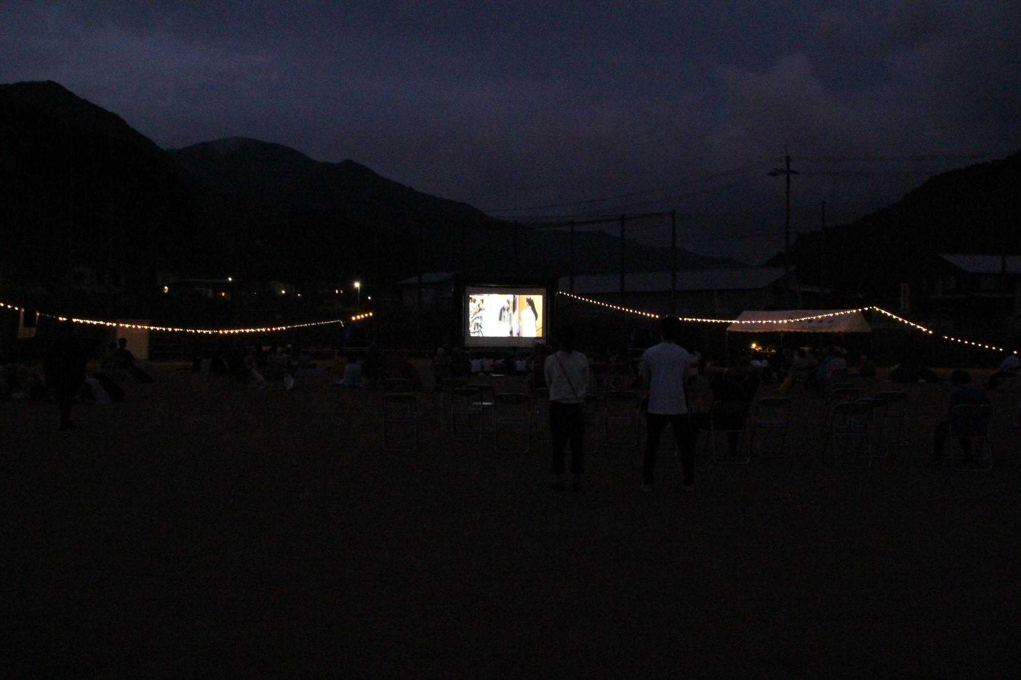 日没後に野外で上映された映画を地域の人が集まって鑑賞している写真