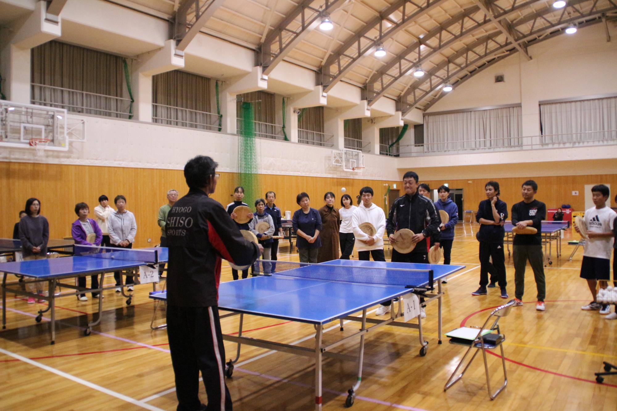 試合の開始前に参加チームのメンバーが集まり、丸太卓球の注意点などをスポーツ推進委員が実演しながら説明している写真