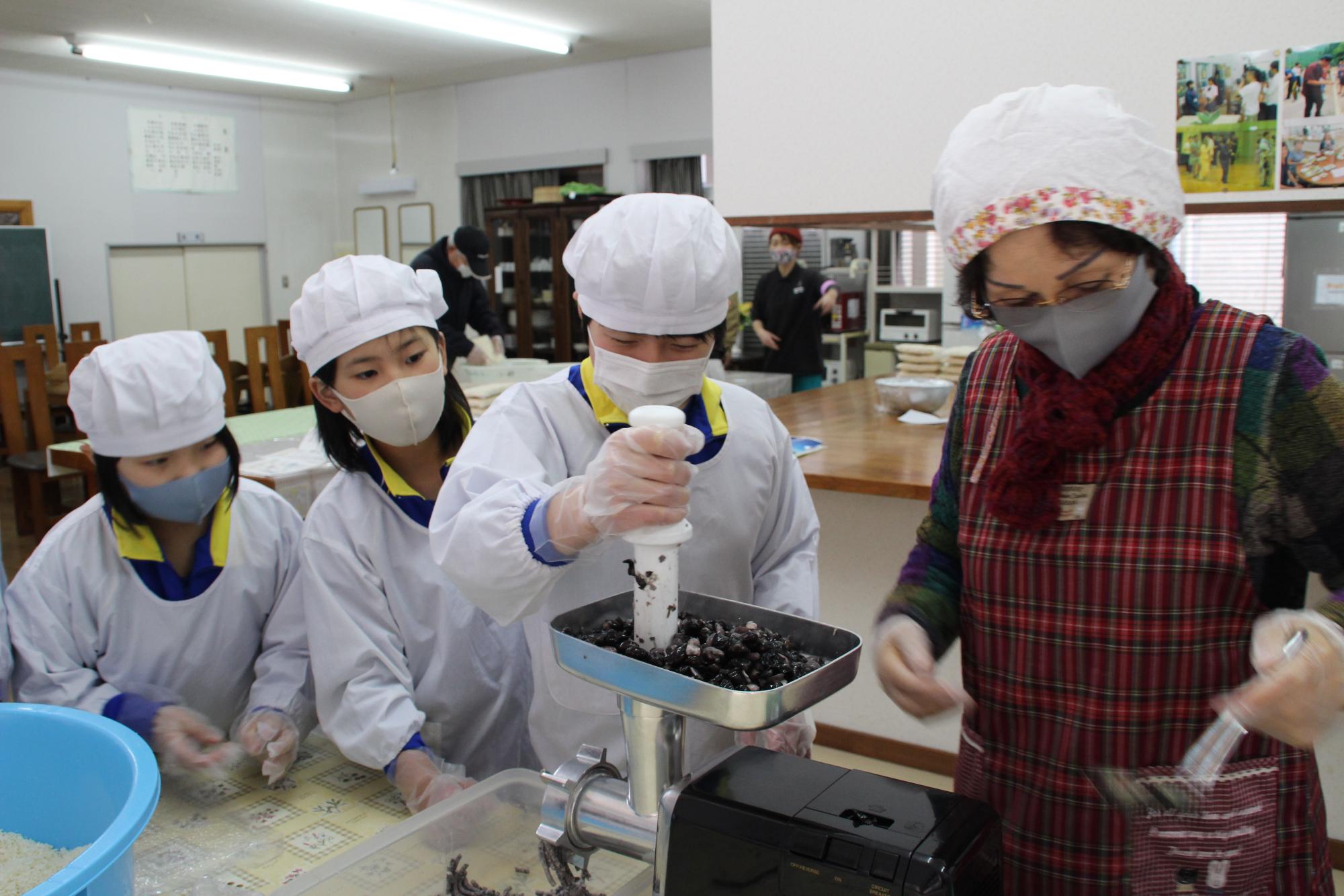 煮た黒大豆をミンサーという機械でつぶしている児童らと指導する女性の写真