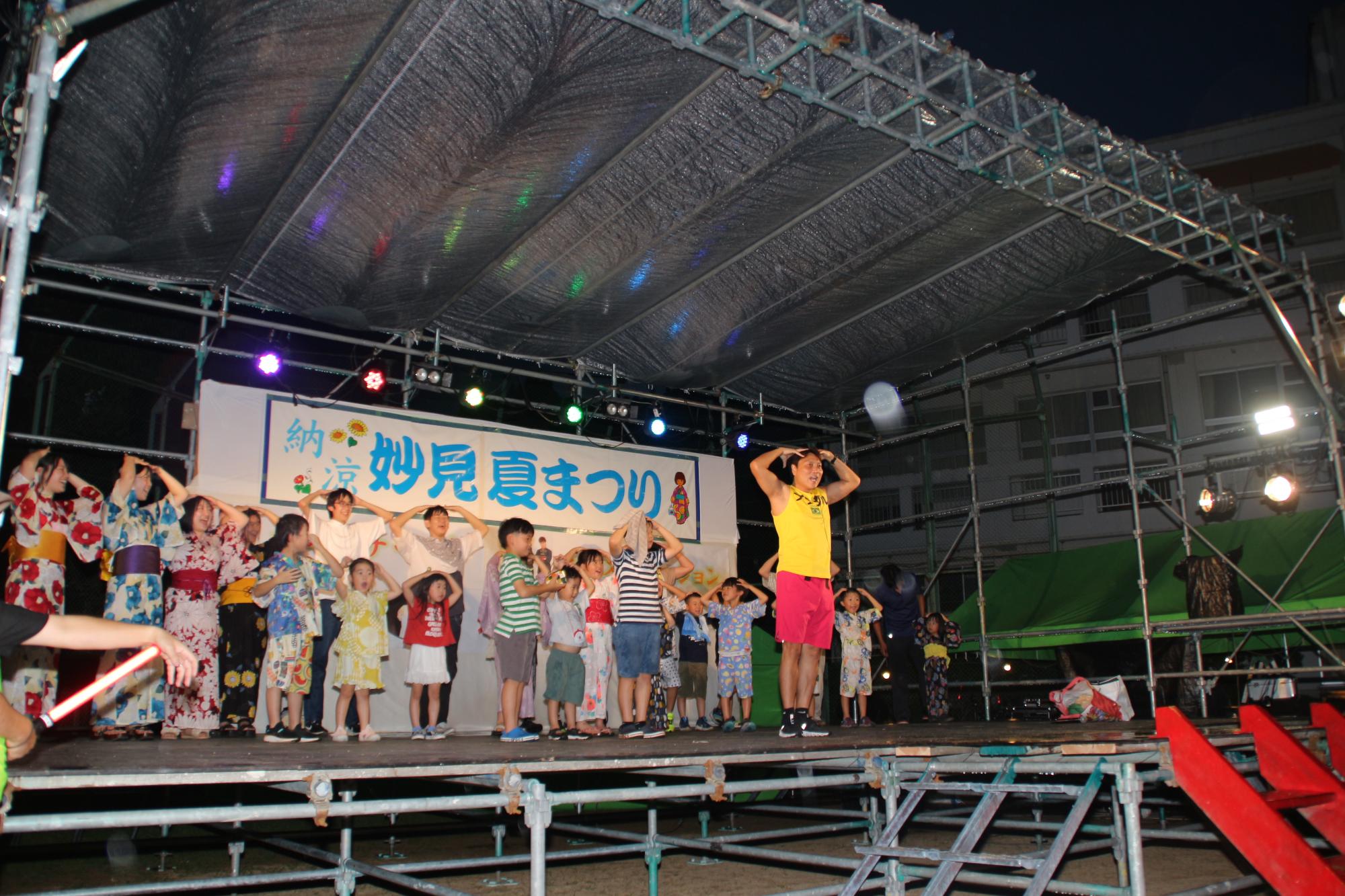 ステージイベントのお笑いライブでサバンナ八木さんが子どもたちをステージに上げ、ギャグを一緒にやっている写真