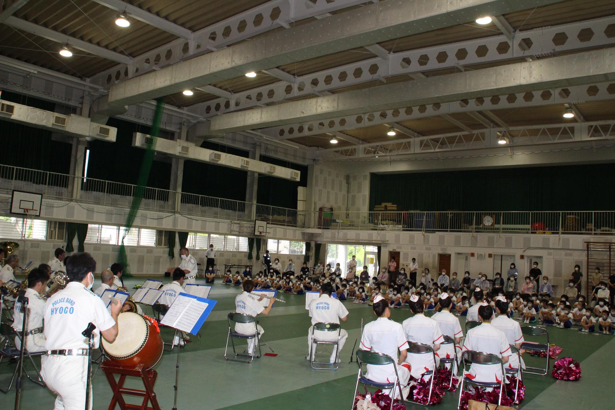 手前に演奏する音楽隊と奥側に演奏を聴く児童らとしきぐさ学園参加者の写真