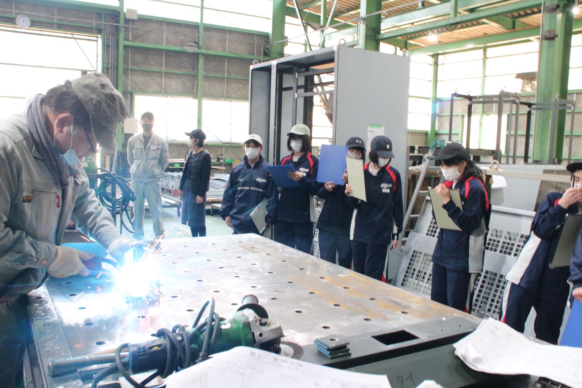 工場内での溶接作業の見学で、飛び散る火花を前に遠巻きに見たり手に持っていた用箋挟で顔を隠したりしながら見学する生徒らの写真