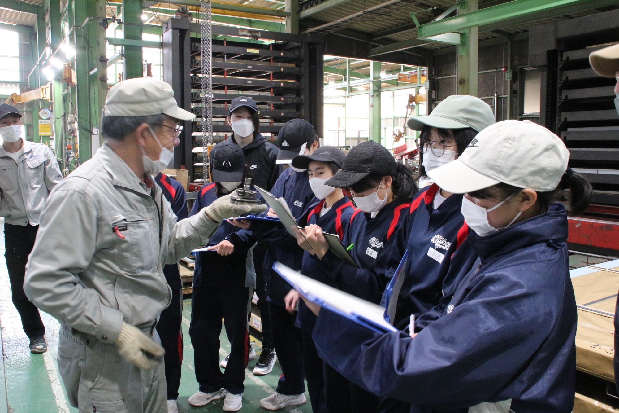 機械の部品を手に持ち、金属に穴を開ける方法を説明する作業員とその周りでメモを取りながら説明を聞いている生徒らの写真