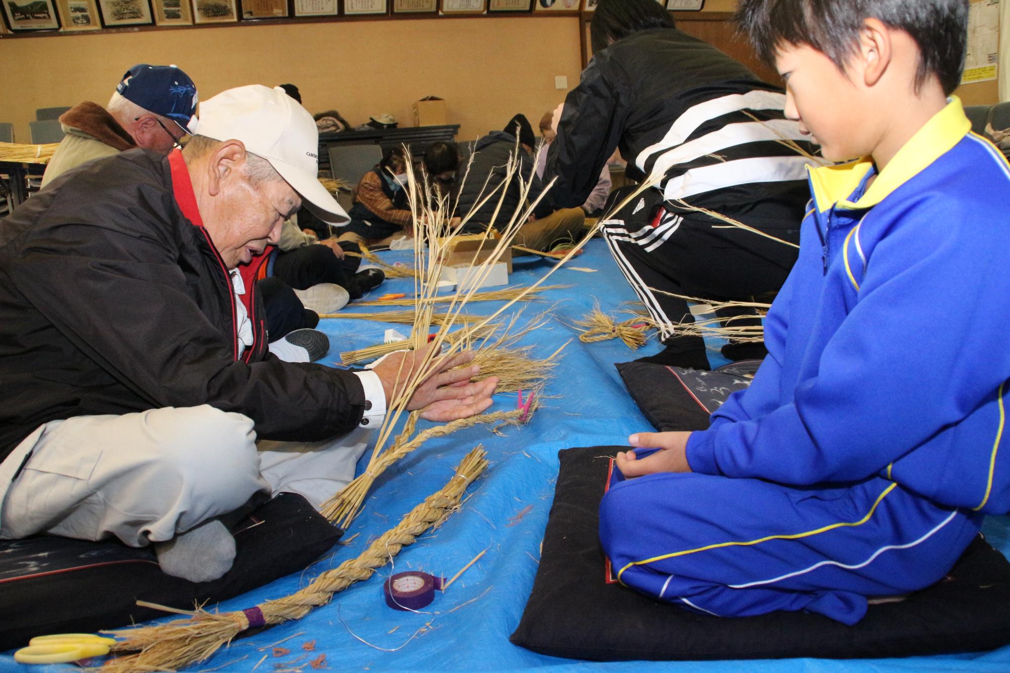 うまくできない低学年の小学生に老人クラブの人が見本を作って見せている写真