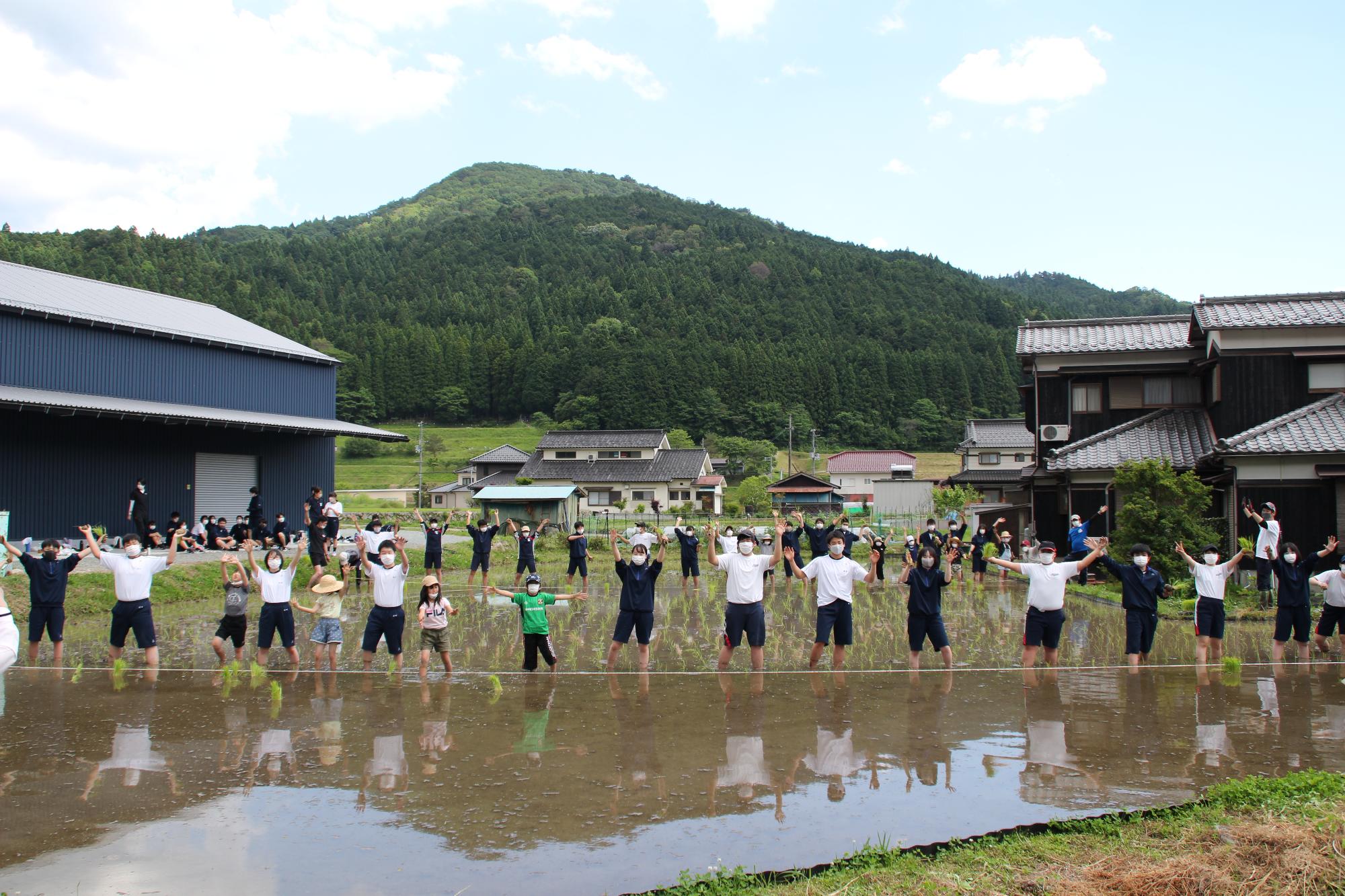 田植えをしている農地の囲んで児童生徒が並び、正面を向いて手を振っている写真