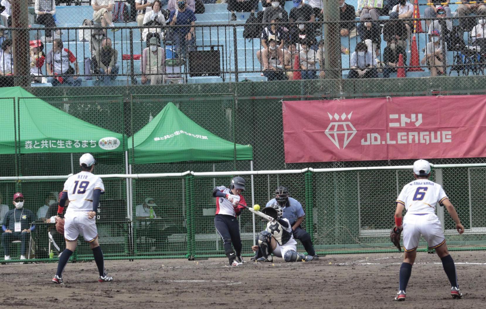 波賀メイプルスタジアムで行われたJDリーグの試合でトヨタ自動織機のピッチャーが投げたボールをシオノギのバッターが打ち返そうとしている写真