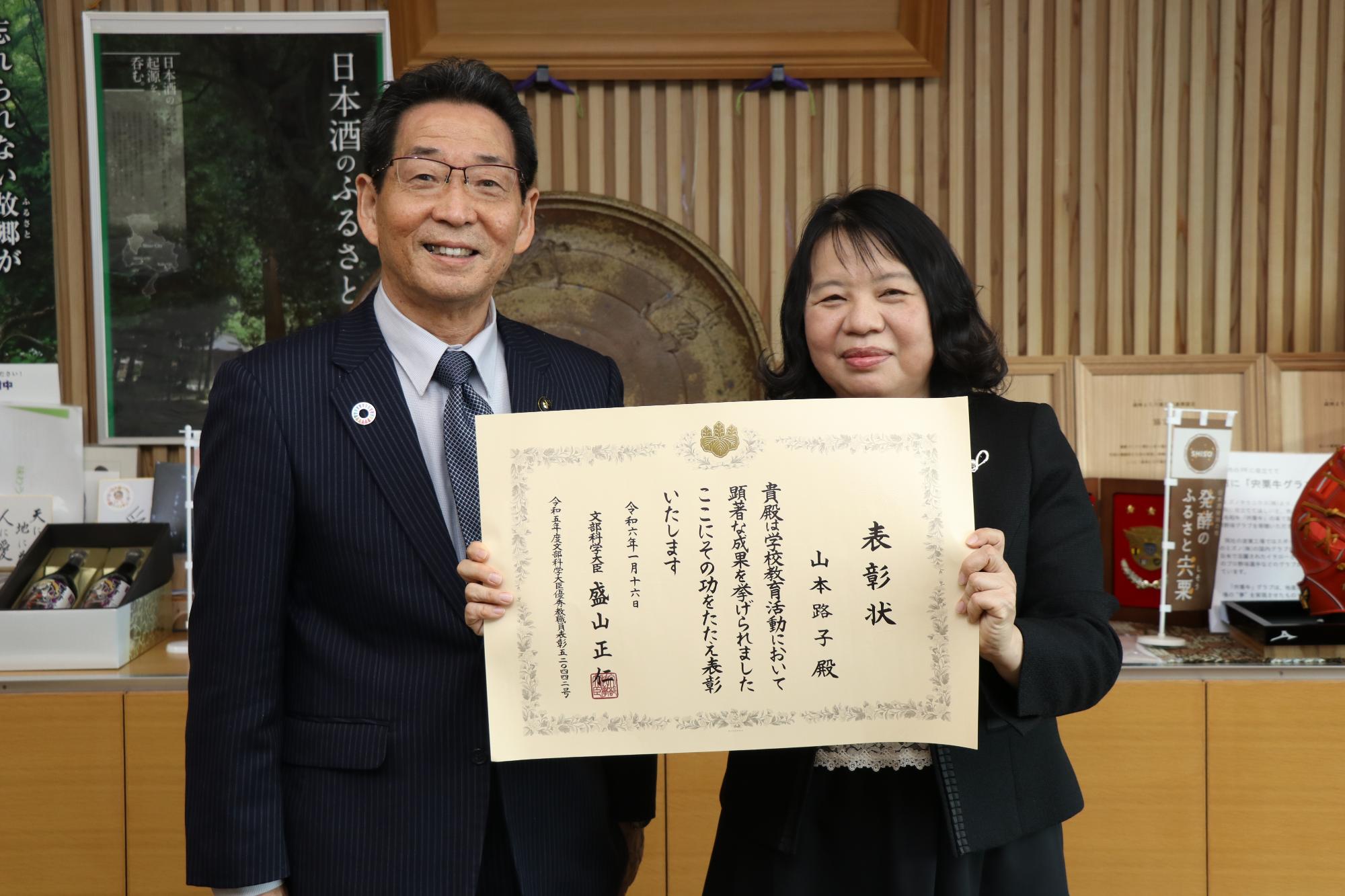 福元市長と文部科学大臣優秀教員表彰の表彰状を手にした戸原小学校の山本養護教諭が並んでいる写真