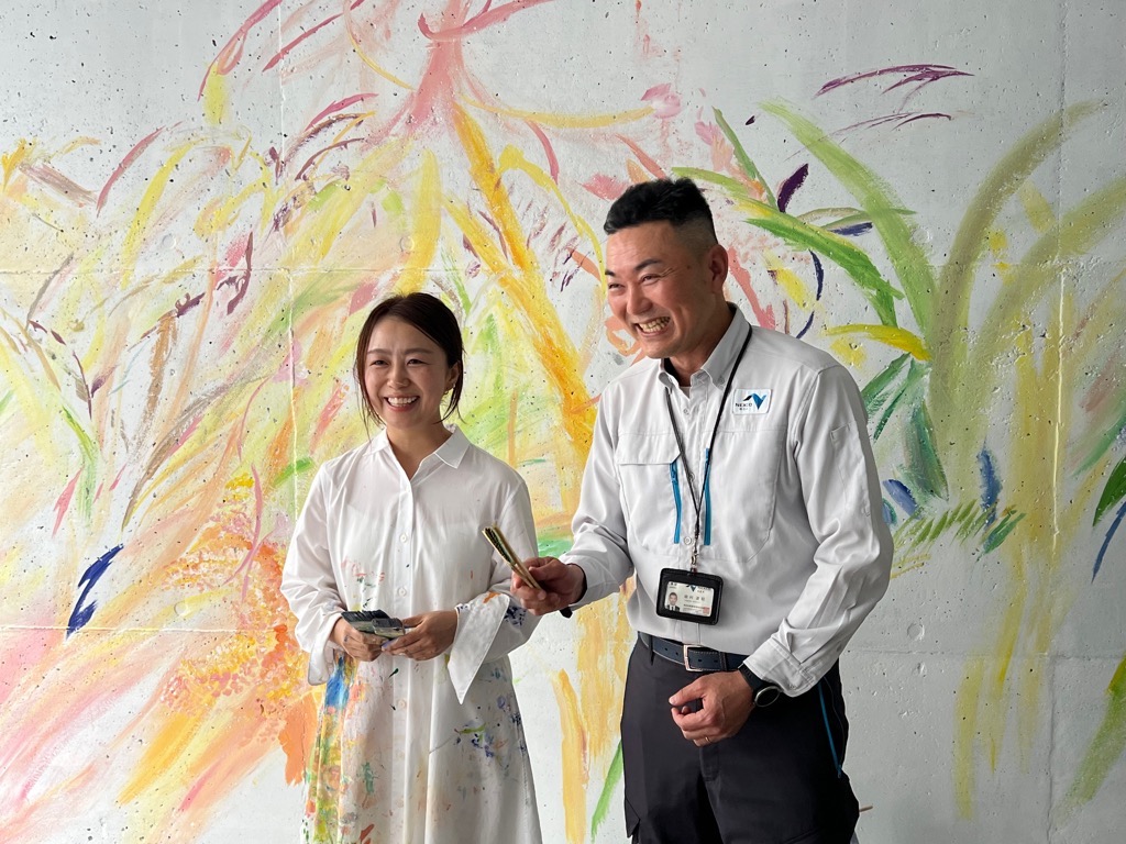 美術作家の植田志保さんとネクスコ西日本福崎道路事務所田村所長が制作中の壁面を背に笑顔で立っている写真