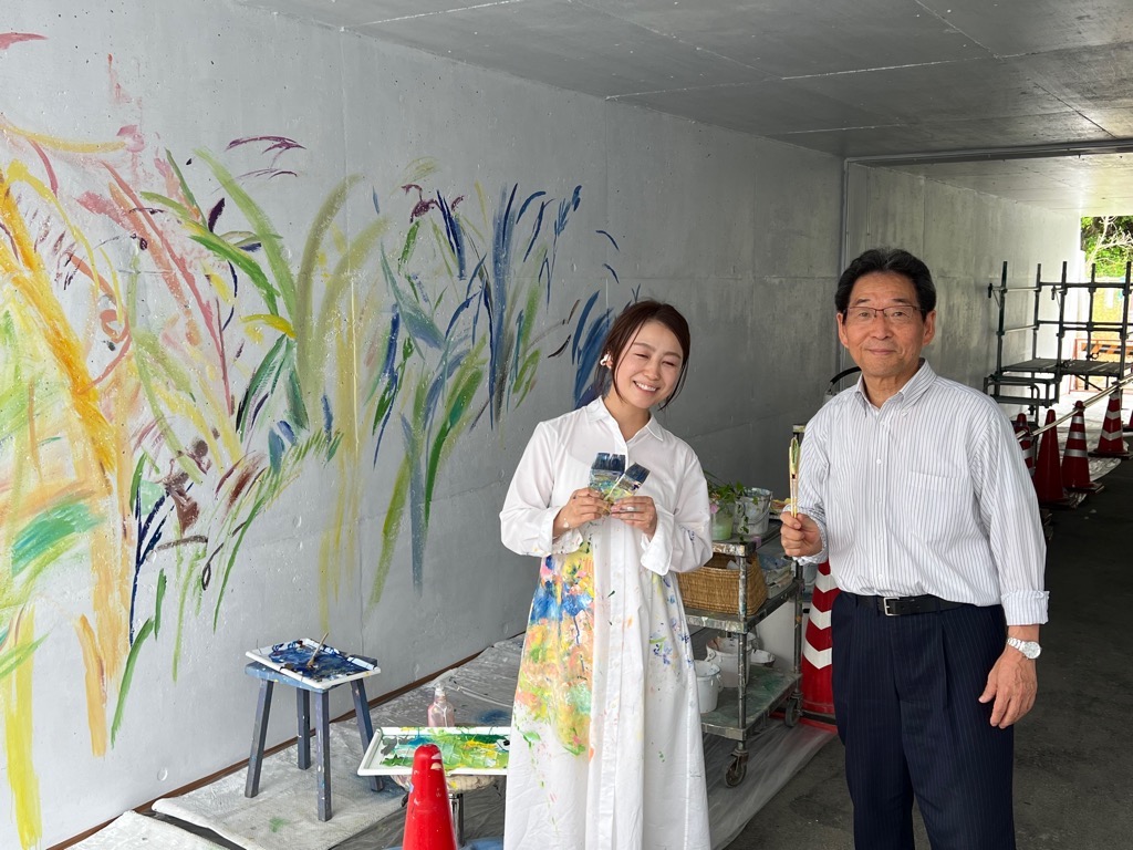美術作家の植田志保さんと福元市長が制作中の通路壁面を背に笑顔で立っている写真